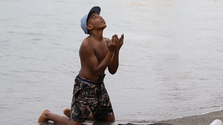 A boy kneeling on the shore of a lake.